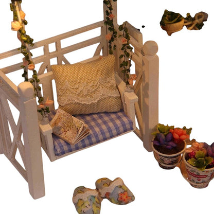COMFORTH miniature dollhouse building kit with a white porch swing, blue gingham cushion, yellow pillow, and potted plants.